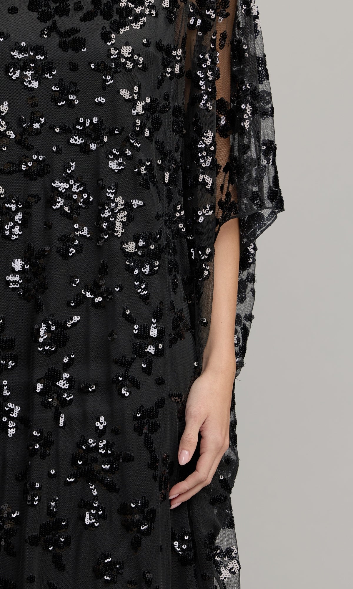 A personโs hand rests by their side, wearing the Mac Duggal 9441 Cape-Sleeve Long Formal Dressโa dramatic black sheer gown adorned with clusters of black and silver sequinsโagainst a plain background.